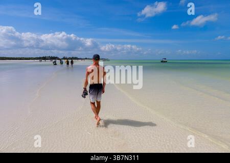 Holbox, Messico - 10 febbraio 2025: Un uomo cammina a piedi nudi lungo il banco di sabbia sulla via del ritorno da Punta Mosquito, incorniciato dalle acque calme e poco profonde e. Foto Stock