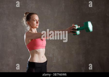 Donna che solleva i manubri in avanti durante l'allenamento di forza in palestra su uno sfondo grigio. Foto Stock