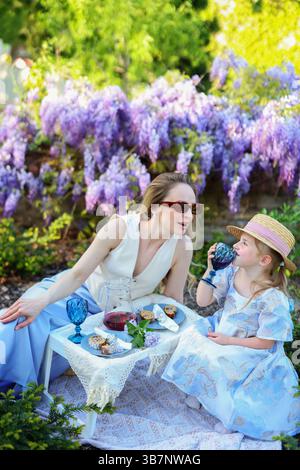 Mamma e ragazza eleganti fanno un picnic con succo di frutta e pasticcini nel cortile soleggiato. Foto Stock
