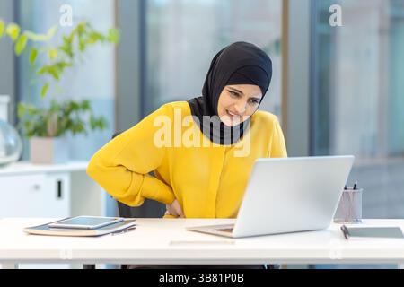 Una donna che indossa un hijab soffre di mal di schiena mentre lavora sul suo notebook alla sua scrivania in un ufficio moderno. Foto Stock
