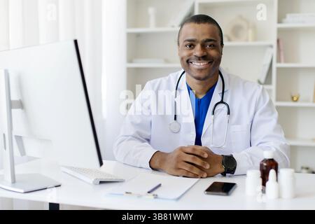 Sorridente bella Black male Doctor in uniforme seduta alla scrivania con computer Foto Stock