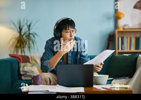 Giovane uomo asiatico che riflette mentre analizza i documenti indossando cuffie con laptop e libri sulla scrivania in un accogliente ambiente domestico Foto Stock