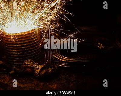 un incredibile primo piano di scintille luminose che volano da un oggetto metallico in un ambiente industriale buio, circondato da cavi a spirale e utensili robusti, catturando Foto Stock