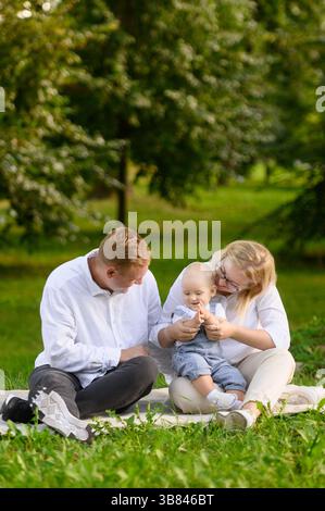 Felice coppia con bambino seduto su una coperta nel parco durante le vacanze, mamma che tiene il bambino in braccio e gioca con lui una torta di pappy, papà felicemente guardando t Foto Stock