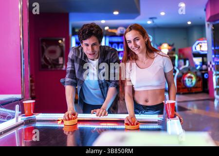 Amici multietnici che trascorrono un fine settimana insieme, giocando a hockey da tavolo in un vivace centro di intrattenimento pieno di giochi arcade e colorate luci al neon Foto Stock