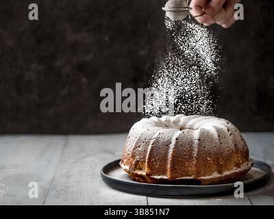 La mano della donna che spruzzava lo zucchero a velo sulla torta di muffin freschi. Lo zucchero in polvere cade su una torta di muffin fresca e perfetta. Spazio di copia per il testo. Idee e ricette per Foto Stock