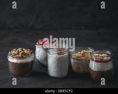Set di budino di chia in diversi vasetti di vetro sul tavolo scuro. Assortimento di chia puding con frutta, noci e ingredienti diversi. Copia spazio per il testo. Su Foto Stock