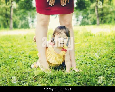 Bambina di un anno seduta tra le gambe della madre sorridente e che guarda la macchina fotografica all'aperto. Figlia che si nasconde tra le gambe della madre sull'erba verde dentro Foto Stock