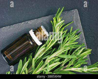 Olio essenziale di rosmarino in vetro scuro e bottiglia di rosmarino fresco su sfondo scuro. Vista superiore o laici piatta Foto Stock