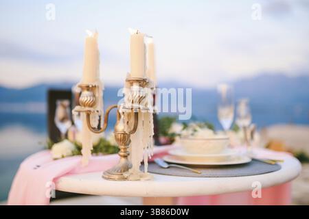 Primo piano di un tavolo da pranzo di nozze al ricevimento. Bruciare le candele in un candelabro su un tavolo con un panno rosa e piatti di crema sullo sfondo di un sole Foto Stock