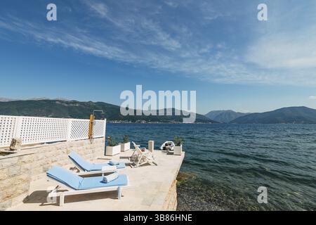 Tivat, Montenegro - 01 luglio 2020: Lettini bianchi in legno sul molo, in riva al mare. Nella baia di Cattaro, in Montenegro. Acqua blu azzurra e fondo ciottolo Foto Stock