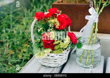 Un cestino di vimini bianco pieno di rose rosse e vegetazione si trova su una panchina resistente agli agenti atmosferici accanto a un vaso con gambi di rose e nastro. Foto Stock