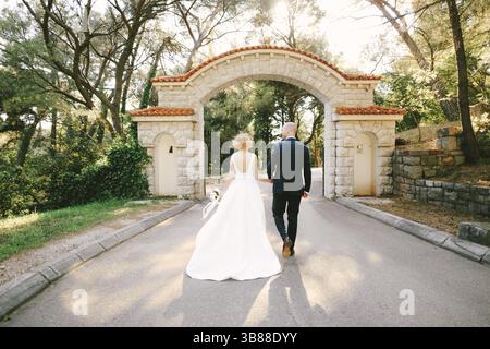 Gli sposi novelli camminano lungo il sentiero tenendo per mano il cancello di pietra dell'ingresso con piastrelle arancioni in un parco verde. Vista posteriore. Foto Stock