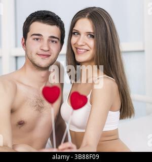 Amore per la coppia di san valentino che tiene il cuore rosso giovane adorabile insieme sdraiato in un letto, sorriso felice guardando la macchina fotografica, cuori concettuali che volano intorno Foto Stock