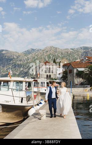 La sposa e lo sposo camminano lungo il molo tenendosi per mano nella baia di Cattaro, barche turistiche vicino al molo . Foto Stock