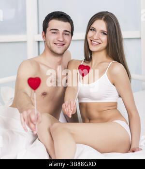 Amore per la coppia di san valentino che tiene il cuore rosso giovane adorabile insieme sdraiato in un letto, sorriso felice guardando la macchina fotografica, cuori concettuali che volano intorno Foto Stock