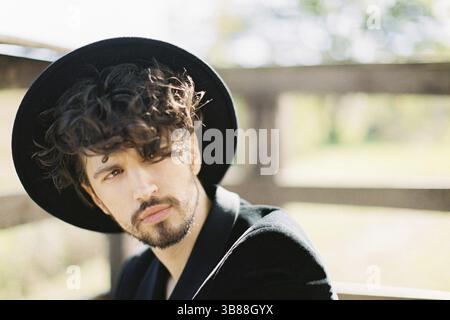 Un giovane con un cappello nero si siede vicino a una ringhiera di legno. Verticale. Foto Stock