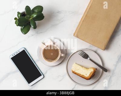 Caffè e cheescake su un piano in marmo bianco. Vista dall'alto o in piano. Tazza da caffè, pezzo di cheesecake, smartphone, libro e succulente. Ufficio domestico, fem Foto Stock