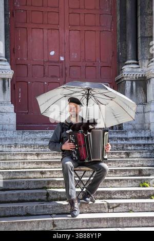 Busker con ombrellone e gatto appoggiato sulla fisarmonica sui gradini della porta laterale Basilique du Sacré-Cœur de Montmartre a Parigi, Francia Foto Stock