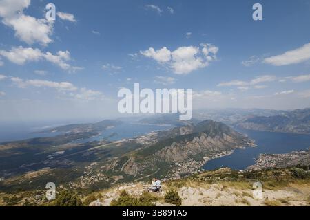 Un uomo e una donna sono seduti in un abbraccio su una panchina, di fronte a loro si apre una vista panoramica della Baia di Cattaro. Filmati FullHD di alta qualità Foto Stock