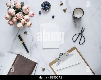 Wedding mock up. Piatto con laici bigliettino vuoto, busta. Appunti, fiori, candele profumate su marmo bianco. Femminile home office mock up con co Foto Stock