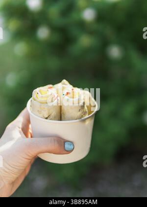 Gelato arrotolato in tazza conica a mano della donna. Donna tiene la tazza del cono con il gelato laminato della banana di kiwi di stile thailandese su sfondo verde delle foglie. Esterno. NAT Foto Stock