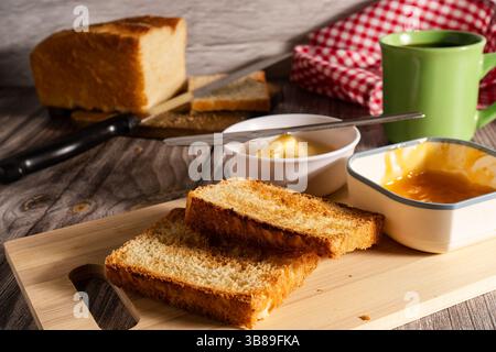 Fette di pane tostato servite su una tavola di legno con burro e marmellata di albicocche, perfette per la colazione Foto Stock