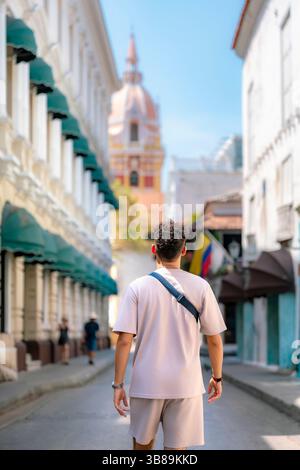 L'inconfondibile giovane uomo latino con capelli ricci e abbigliamento beige informale esplora la storica via Cartagena con la torre della cattedrale e la bandiera colombiana. Foto Stock