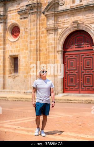 Sorridente giovane latino in abiti leggeri e casual e cappello di paglia in piedi di fronte alla storica chiesa in pietra con porta rossa a Cartagena, Colombia. Foto Stock