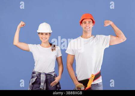 Uomo allegro e donna in caschi protettivi che alzano i pugni, mostrando i muscoli bicipiti Foto Stock