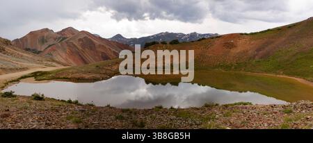 I cieli ricoperti di nuvole si stagliano su un tranquillo laghetto di montagna, che riflette le vette frastagliate e i vari toni della terra del paesaggio del Colorado. I sereni setti Foto Stock