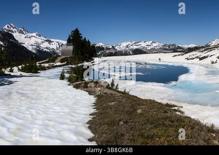La neve ricopre il terreno mentre un tranquillo lago glaciale riflette il cielo azzurro. I pini punteggiano il paesaggio tra torreggianti montagne, creando uno stupendo panorama Foto Stock