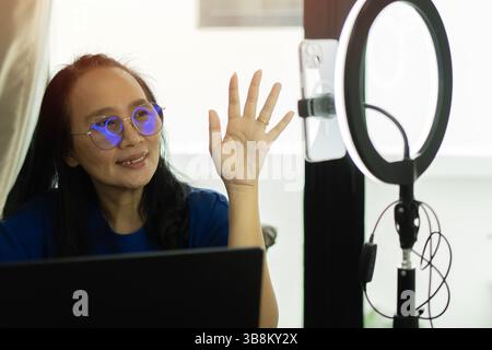 Donna asiatica felice che lavora a casa con computer portatile, telefono cellulare e illuminatore anulare che produce contenuti per il canale web. Foto Stock