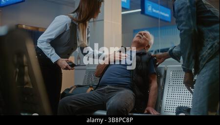 Affollato terminal dell'aeroporto internazionale: Uomo anziano infarto, torace in dolore seduto nella sala partenze dell'hub aereo. Persone diverse e personale aeroportuale che aiuta i passeggeri anziani. Foto Stock
