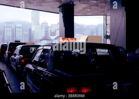 La coda dei taxi sotto la tettoia geometrica con lo skyline di Hong Kong sullo sfondo Foto Stock
