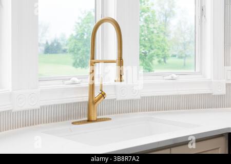 Un rubinetto da cucina dorato su un ripiano in marmo bianco, piastrelle abbronzanti, armadi marroni e finestre rivestite di bianco. Foto Stock