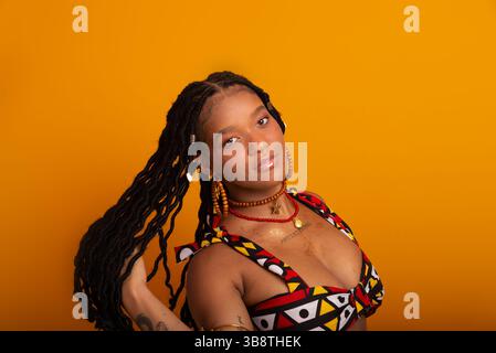 Bella giovane donna con i capelli intrecciati che indossa abiti tradizionali, in posa per la fotocamera. Isolato su sfondo color senape. Sicuro di sé e cal Foto Stock
