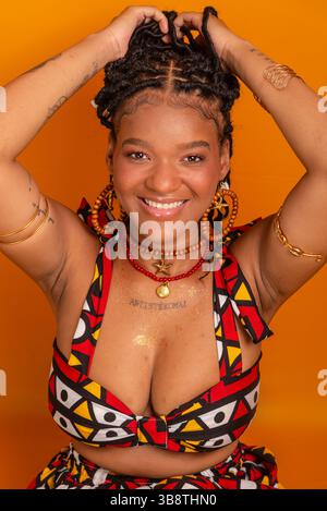 Bella giovane donna seduta con trecce tra i capelli che indossa abiti tradizionali e posa per la fotocamera. Isolato su sfondo color senape. Con Foto Stock