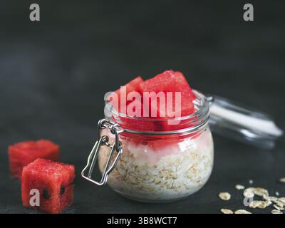 Colazione sana: Avena notturna con pezzi di anguria in vaso di vetro. Porridge di farinata d'avena con anguria. Avena notturna su sfondo nero. Foto Stock