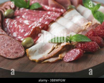 Piatto di antipasti su un piatto di legno da vicino. Piatto di carne affumicata fredda con salsiccia, prosciutto a fette, prosciutto, pancetta, olive e basilico. Antipasto su woo Foto Stock