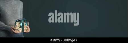Donna in pullover grigio in cashmere che regge una luminosa confezione regalo con le mani su sfondo verde scuro. Lampadine gialle incandescenti in una confezione regalo trasparente con Foto Stock