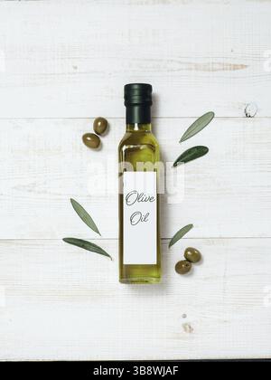 Modello della bottiglia di vetro dell'olio d'oliva. Vista dall'alto della bottiglia di vetro verde con olio extra vergine d'oliva su sfondo di legno bianco con olive verdi e gree fresco Foto Stock