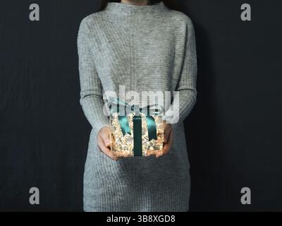 Donna in pullover grigio che tiene in mano una scatola regalo luminosa su sfondo scuro. Lampadine gialle brillanti in una confezione regalo trasparente con nervature di colore verde scuro Foto Stock