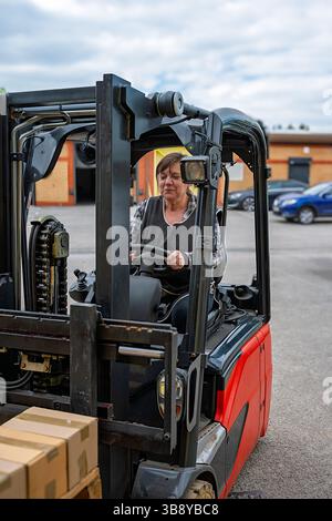 Una donna sta guidando un carrello elevatore. Il carrello è rosso e nero. Ci sono delle scatole a terra Foto Stock