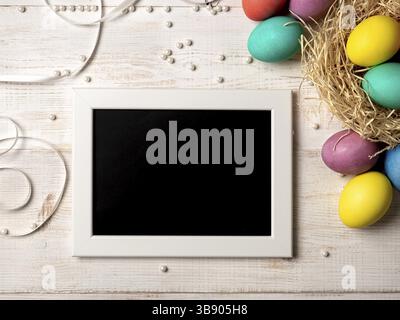 Concetto di Pasqua. Uova colorate su sfondo di legno bianco con lavagna vuota. Copiare spazio per messaggi di saluto, testo o design. Vista dall'alto verso il basso o piatto Foto Stock