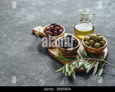 Set di olive verdi, nere e rosse o rosa e olio d'oliva su sfondo grigio. Diversi tipi di olive in ciotole di legno d'oliva e olio d'oliva su legno Foto Stock