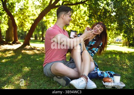 Una giovane coppia felice sta facendo un picnic sull'erba in un parco soleggiato, gustando spuntini e caffè insieme. Quell'uomo sta offrendo una pasticceria al riso Foto Stock