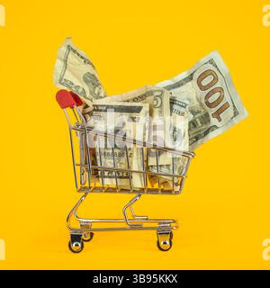 Carrello della spesa pieno di soldi in banconote in dollari USA su sfondo giallo. Simboleggiando l'inflazione o l'aumento del costo della vita e l'aumento del costo delle basi Foto Stock