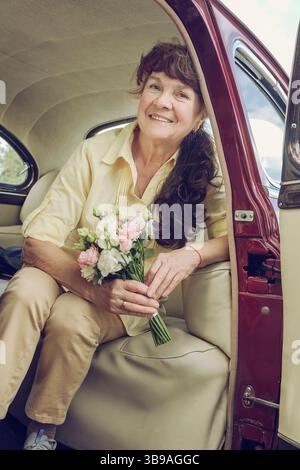 una donna anziana sorridente con un bouquet di fiori è seduta nella cabina di un'auto d'epoca Foto Stock