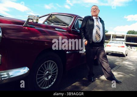 un uomo anziano vestito con una giacca nera su una strada in città in un'auto d'epoca Foto Stock